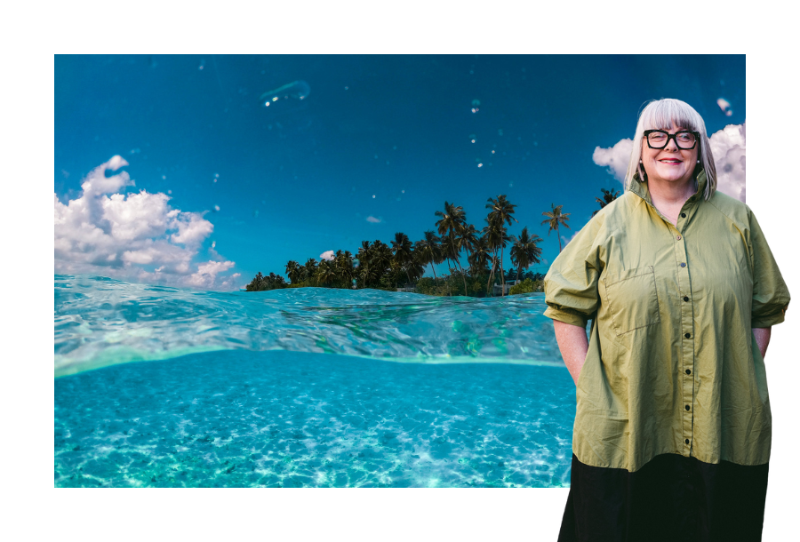 Pictured, Anthea Taylor, standing in front of a picture of a beautiful blue cleared water beach and sandy shores. There are palm trees in the background and a crystal clear blue sky.