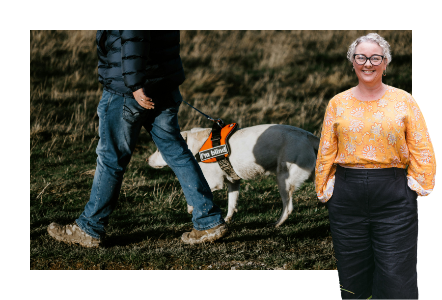 Pictured, Amy Osborne with her hands in her pockets and a bright yellow and orange coloured top. A big smile on her face and in the background is a person walking with their dog. The white dog is wearing a lead that says "I'm Blind" indicating it's ability to help with low vision people.