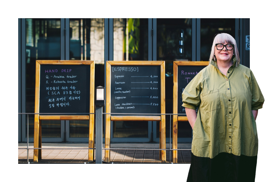 Pictured: Anthea Taylor standing smiling. Behind them is an image of three chalk menu boards out the front of a cafe.