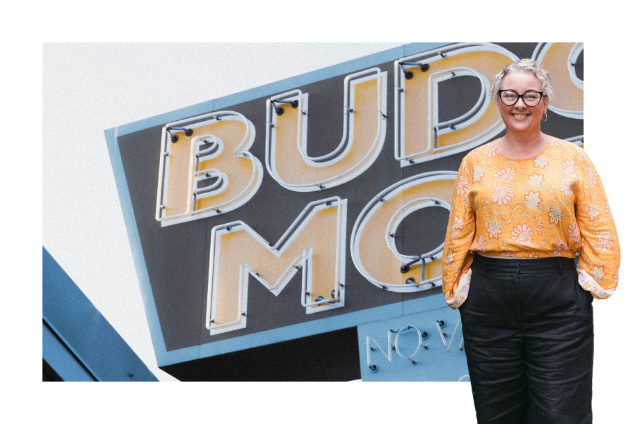 Pictured, Amy Osborne, standing in an orange long sleeve top and black trousers. Behind her is an image of a motel sign.