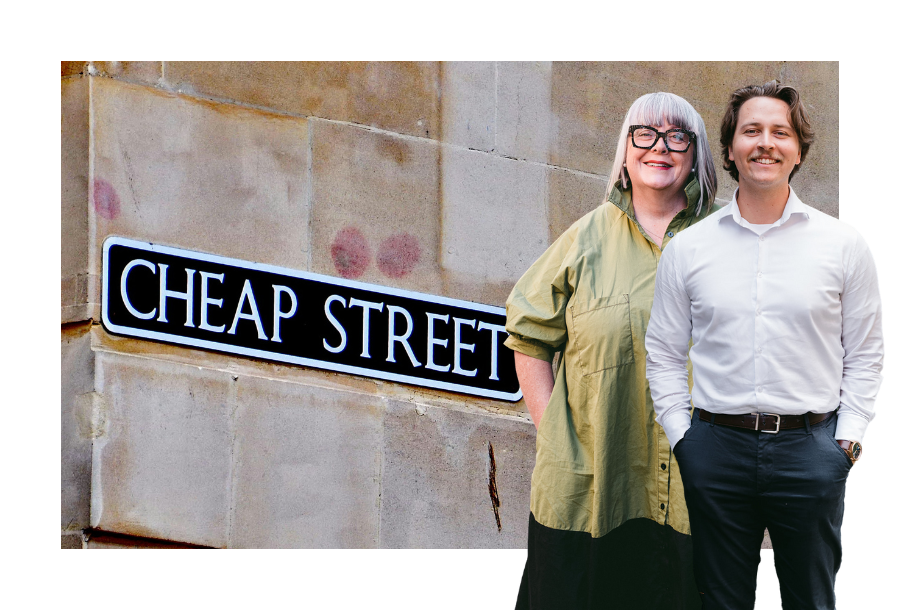 Pictured, Zachary Henderson standing smiling in white business button up and navy-blue trousers and Anthea Taylor standing behind him. Behind them is an image of a can of some kind with a special sticker on it, denoting it's price as 1 dollar.